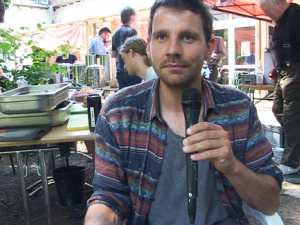Tobi Rosswog mit Mikrophon in der Hand sitzt in einem Innenhof vor einem Tisch, auf dem Essen gemacht wird